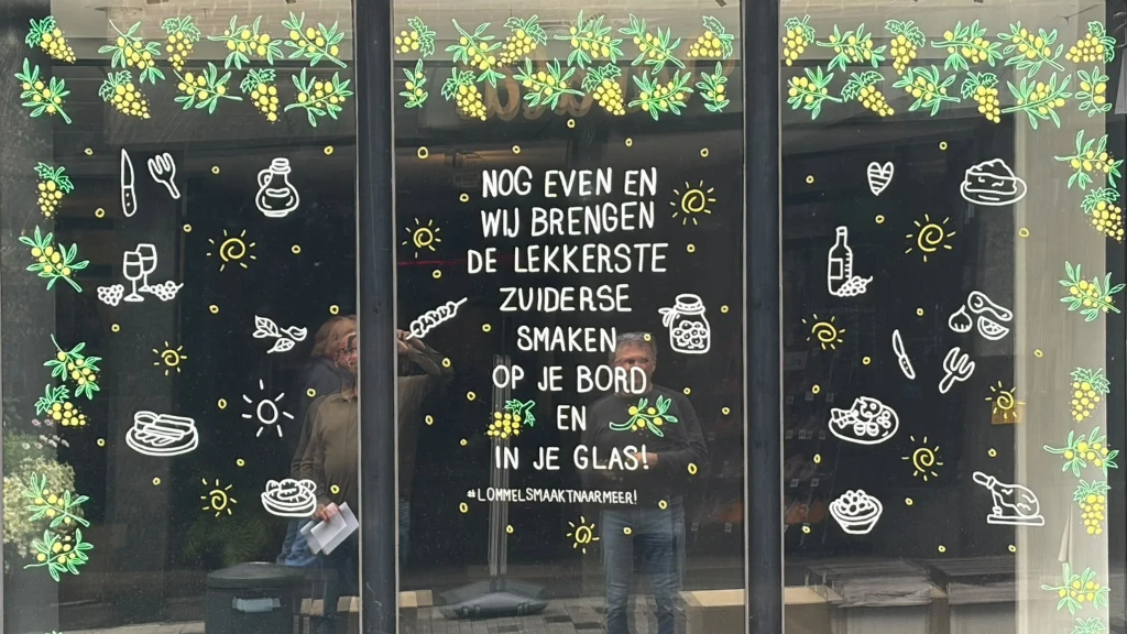 Etalageraam versierd met illustraties van olijftakken en druiventrossen en de tekst nog even en wij brengen de lekkerste zuiderse smaken op je bord en in je glas, met de hashtag lommelsmaaktnaarmeer